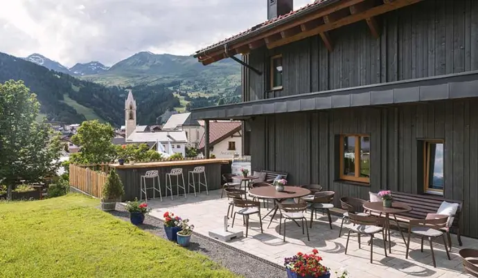 Terrasse mit Tischen vor einem dunklen Holzhaus, mit Blick auf Berge und einen Kirchturm.