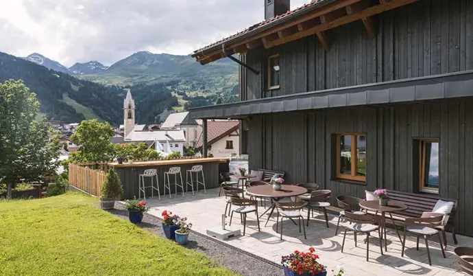 Terrasse mit Tischen vor einem dunklen Holzhaus, Berglandschaft und Kirchturm im Hintergrund. in Pfunds